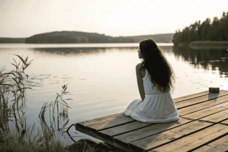 A woman embodying feminine energy sits peacefully on a dock watching the sunrise over a calm lake, inviting a moment of stillness.